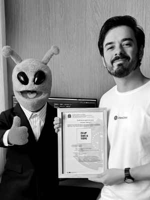O personagem Etefânio, um alienígena verde de terno, ao lado de José Brognoli segurando o certificado da marca Eu Bati na Nave